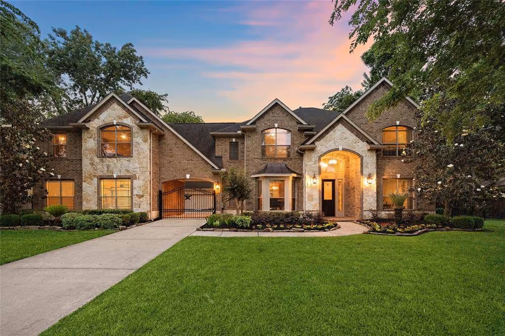 Two-story brick and stone home at dusk. Warm light fills its windows, and lanterns glow beside the stone entry arch and its dark front door. A wrought-iron gate with a star closes the opening at the end of the driveway. Stone-edged flower beds and a mown lawn fill the foreground under a pink and blue sky, with tall trees on either side.